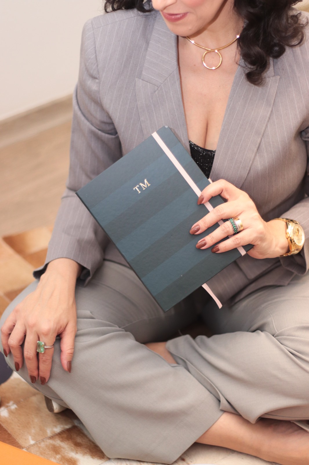 Tati sentada, de blazer cinza, segurando o caderno com o monograma TM